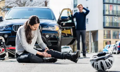 Woman holding her injured leg beside a car accident.